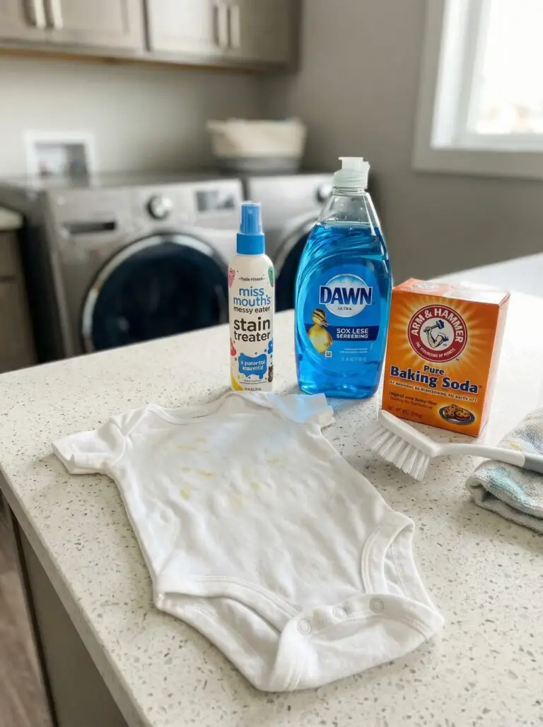 A stained white baby onesie on a white laundry room counter surrounded by stain-fighting supplies, including Dawn dish soap, Arm & Hammer baking soda, Miss Mouth’s Messy Eater spray, and a scrub brush.