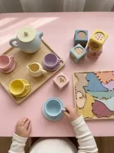 Toddler playing with Montessori silicone toys including Baby Bertie tea set, counting blocks, and dinosaur puzzle on pink play table