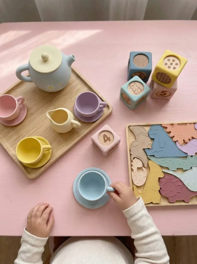 Toddler playing with Montessori silicone toys including Baby Bertie tea set, counting blocks, and dinosaur puzzle on pink play table