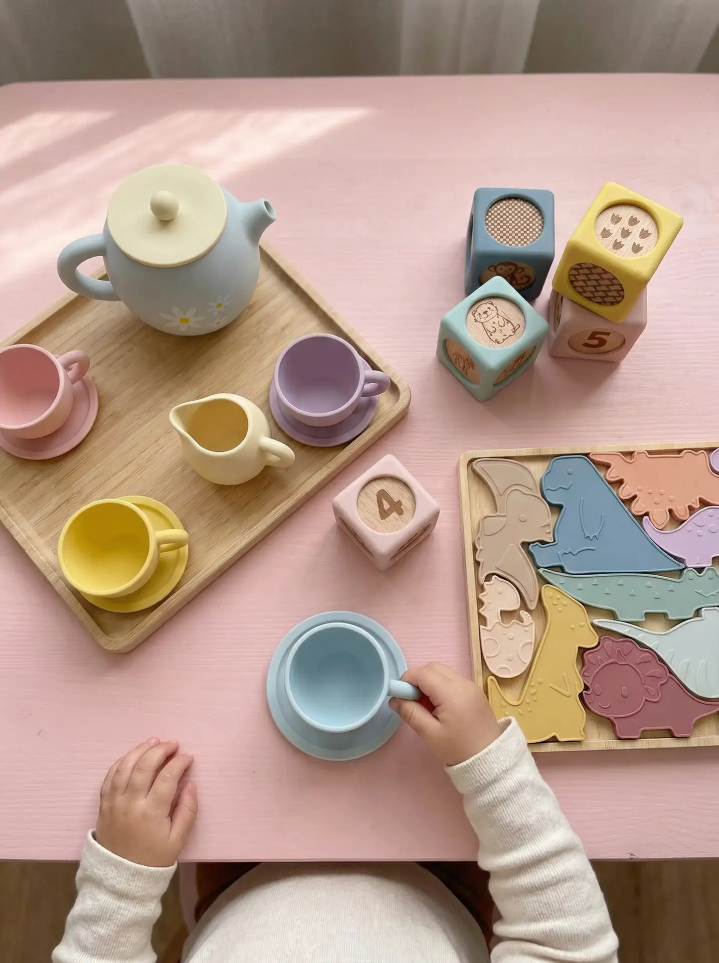 Toddler playing with Montessori silicone toys including Baby Bertie tea set, counting blocks, and dinosaur puzzle on pink play table