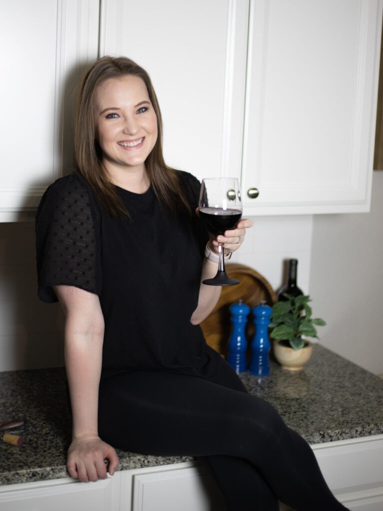 Smiling woman in a black dress sits on a kitchen counter, holding a glass of red wine.